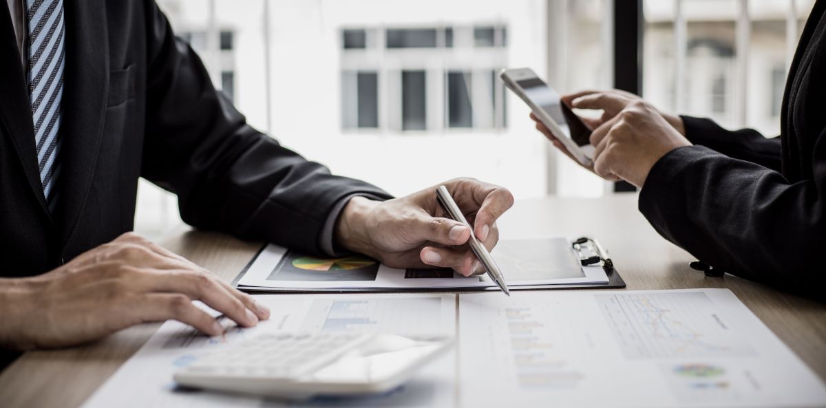 Two business people work in a conference room, one with a calculator and pen, the other with a smartphone.